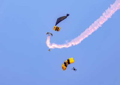 Four skydivers with yellow parachutes and smoke trails perform stunts against a clear blue sky.