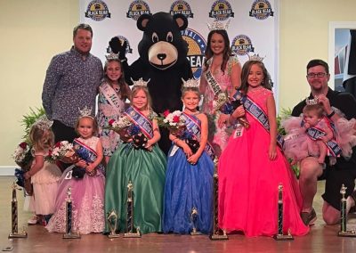 Children in pageant dresses and adults pose with trophies and a bear mascot at a beauty pageant event.