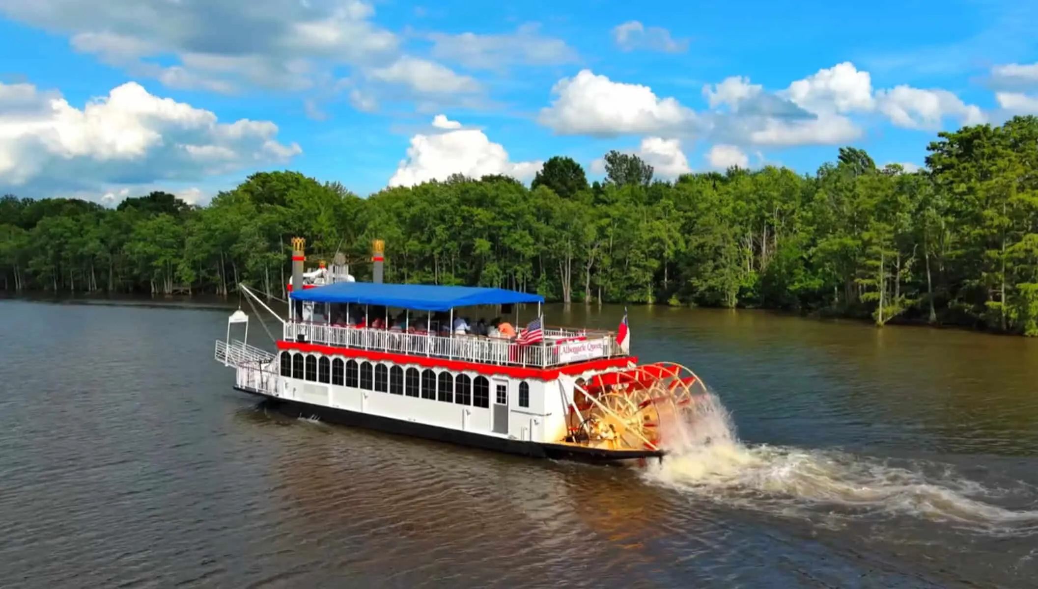 A white and red paddlewheel riverboat cruises along a river with trees and blue sky in the background.