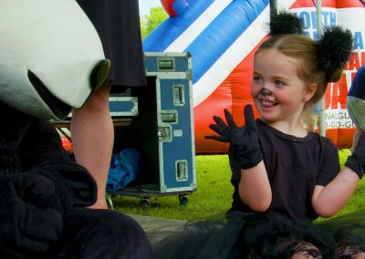 Young girl in a black cat costume smiles and waves at a person in a large animal costume near an inflatable slide.