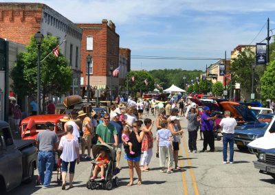 People walk along a busy small-town street fair with classic cars on display and flags hanging from buildings.