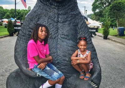 Two children pose with a large bear statue outdoors at a park or festival, smiling at the camera.