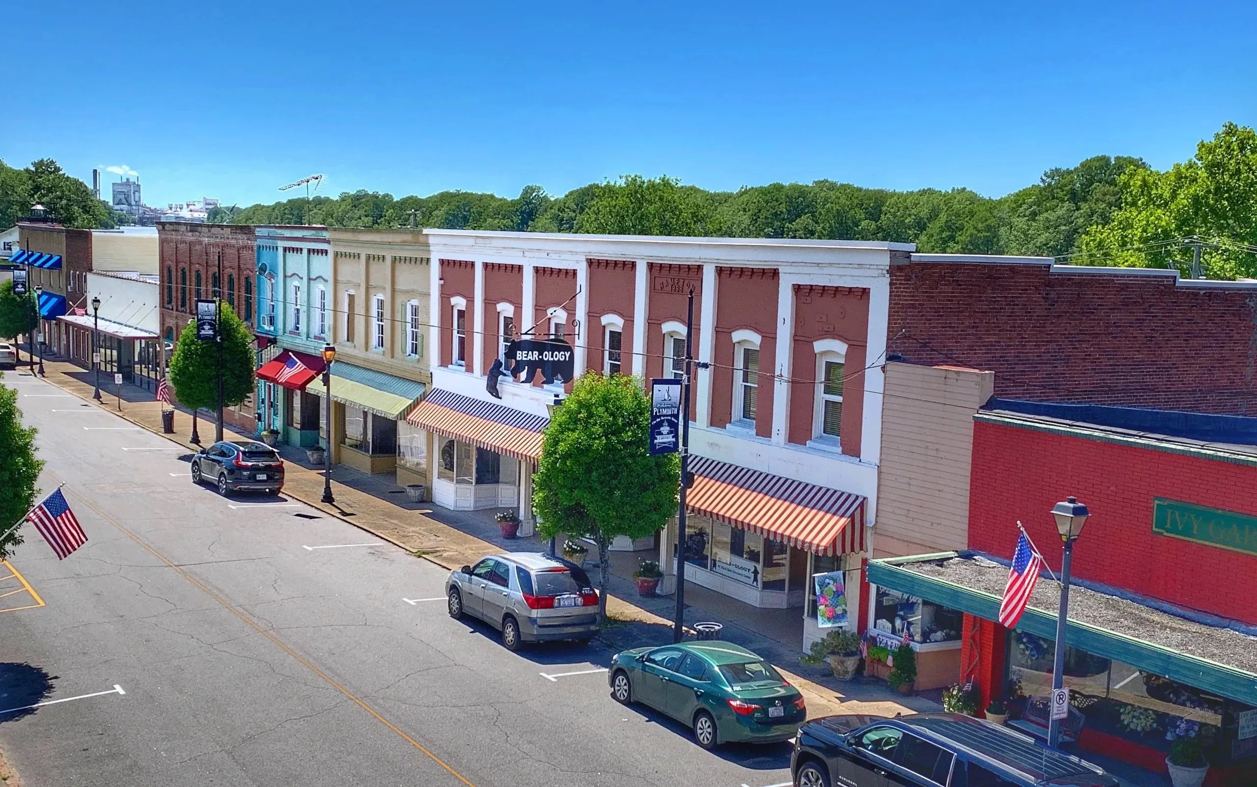 Colorful storefronts with awnings line a small town street with parked cars and trees under a clear blue sky.