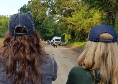 Two people watch a bear crossing a dirt road as others gather near a white van in the distance.