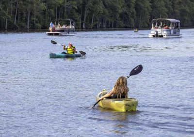 A woman kayaks on a river toward other kayakers and boats, with trees lining the riverbank in the background.