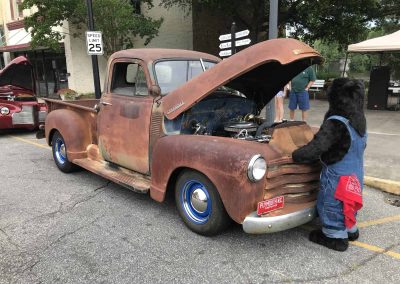 A person in a bear costume examines the engine of a rusty vintage pickup truck at a car show.