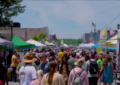 Crowd walking through a busy outdoor street market with tents, flags, and food stalls on a sunny day.