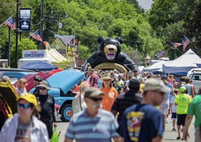 Crowd at an outdoor festival with a giant inflatable black bear and classic cars on display.