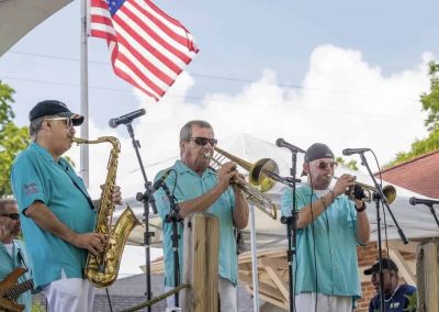 Three men in blue shirts play saxophone, trombone, and trumpet onstage outdoors, with a US flag in the background.