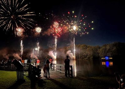 People watch colorful fireworks over a lake at night, standing on a dock near trees and a “Bear Town” sign.
