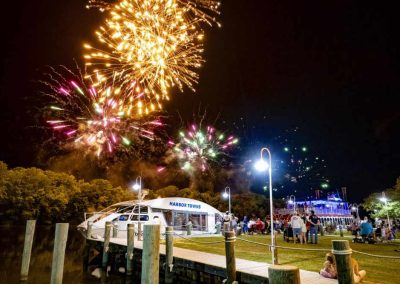 Fireworks light up the night sky over a dock with a boat and people gathered near the water.