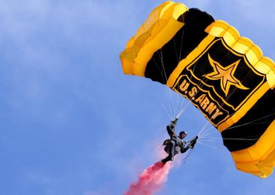 Parachutist descending with a yellow U.S. Army parachute against blue sky, trailing red smoke.