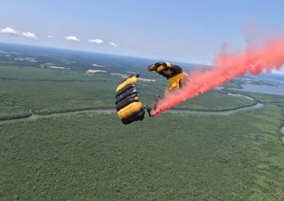 Skydiver with a yellow parachute releases red smoke above a green landscape with rivers and blue sky.