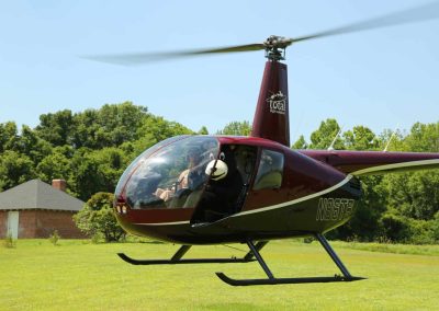 A maroon and white helicopter hovers low over a grassy field near a small house and trees.