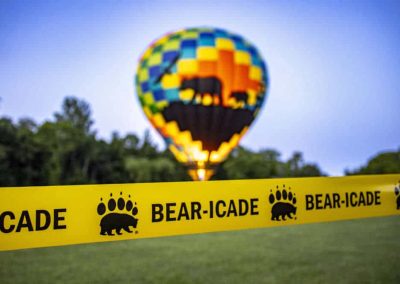 A colorful hot air balloon with a bear design is behind yellow tape labeled "BEAR-ICADE" on a grassy field.