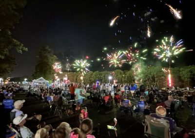 Crowd watching colorful fireworks in the night sky at an outdoor event, seated on lawn chairs.