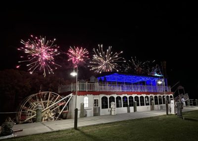 Fireworks light up the night sky above a brightly lit riverboat docked by a grassy shore.
