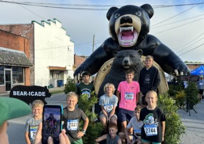 Group of kids pose in front of a large inflatable bear at an outdoor event while someone takes their photo.