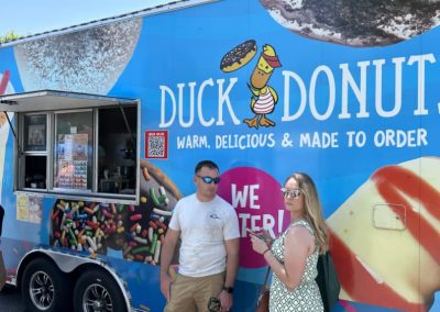 People stand by a colorful Duck Donuts food truck with large donut images and a service window.