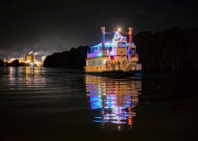 A brightly lit paddlewheel riverboat reflects on calm water at night, with a lit industrial area in the background.