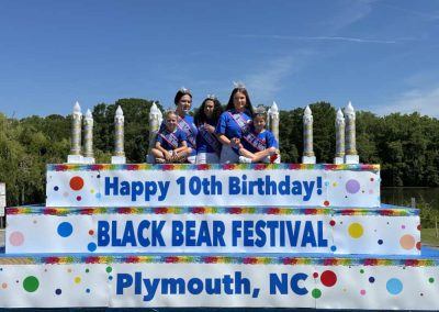Six girls in pageant sashes and crowns on a cake float for the Black Bear Festival in Plymouth, NC.