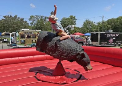 Person riding a mechanical bear at an outdoor event with food trucks and tents in the background.
