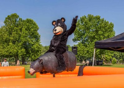 Person in a bear costume rides a mechanical bull outdoors, surrounded by an orange inflatable barrier.