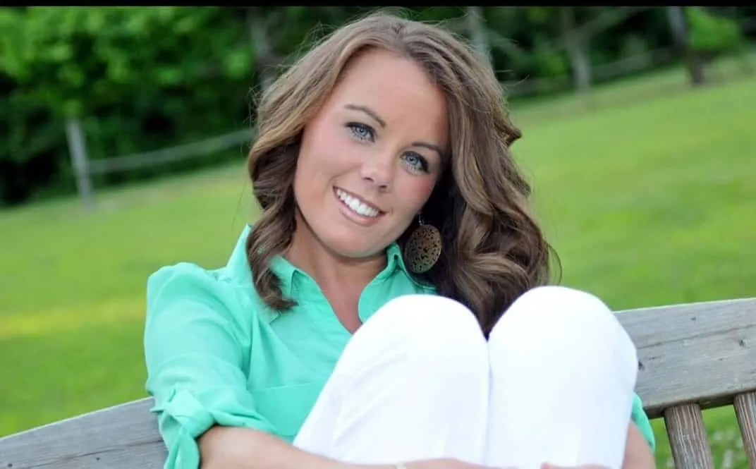 Woman with wavy brown hair wearing a green shirt and white pants, smiling while sitting on a wooden bench outdoors.