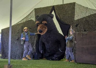 Two people guide a large, realistic bear costume out of a camouflage tent at an outdoor event.