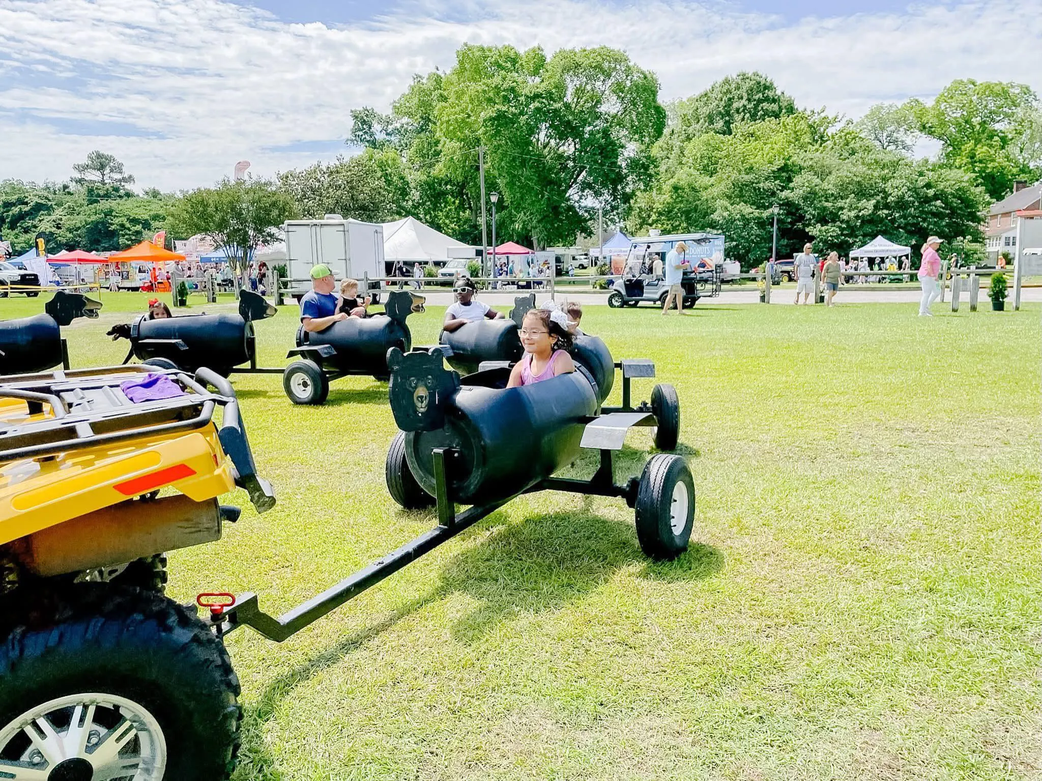 Children ride in black barrel cars pulled by an ATV at an outdoor fair on a sunny day.