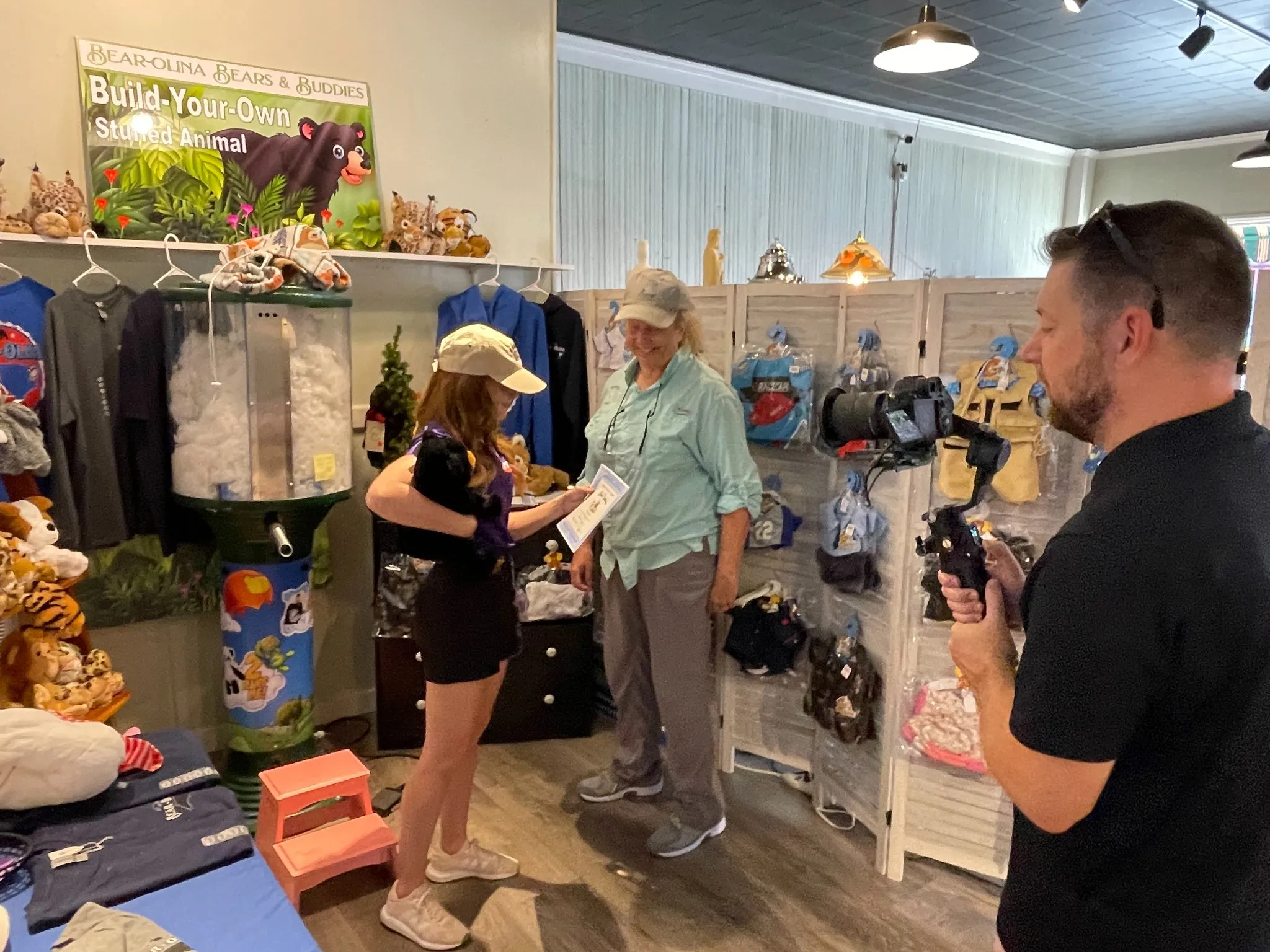 Two women talk in a gift shop with plush toys as a man films them using a camera on a handheld stabilizer.
