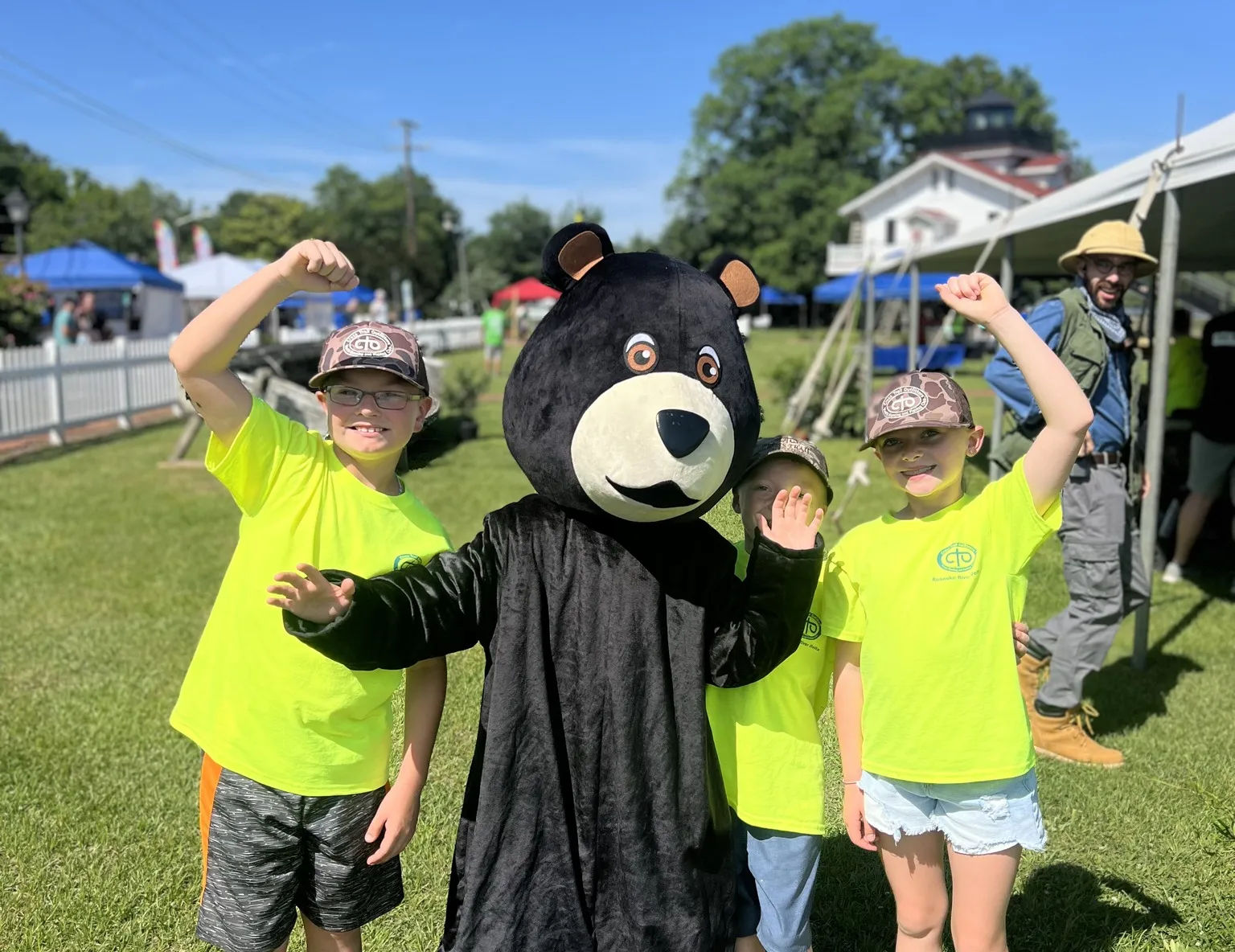 Three kids in yellow shirts pose with a person in a bear costume at an outdoor event on a sunny day.