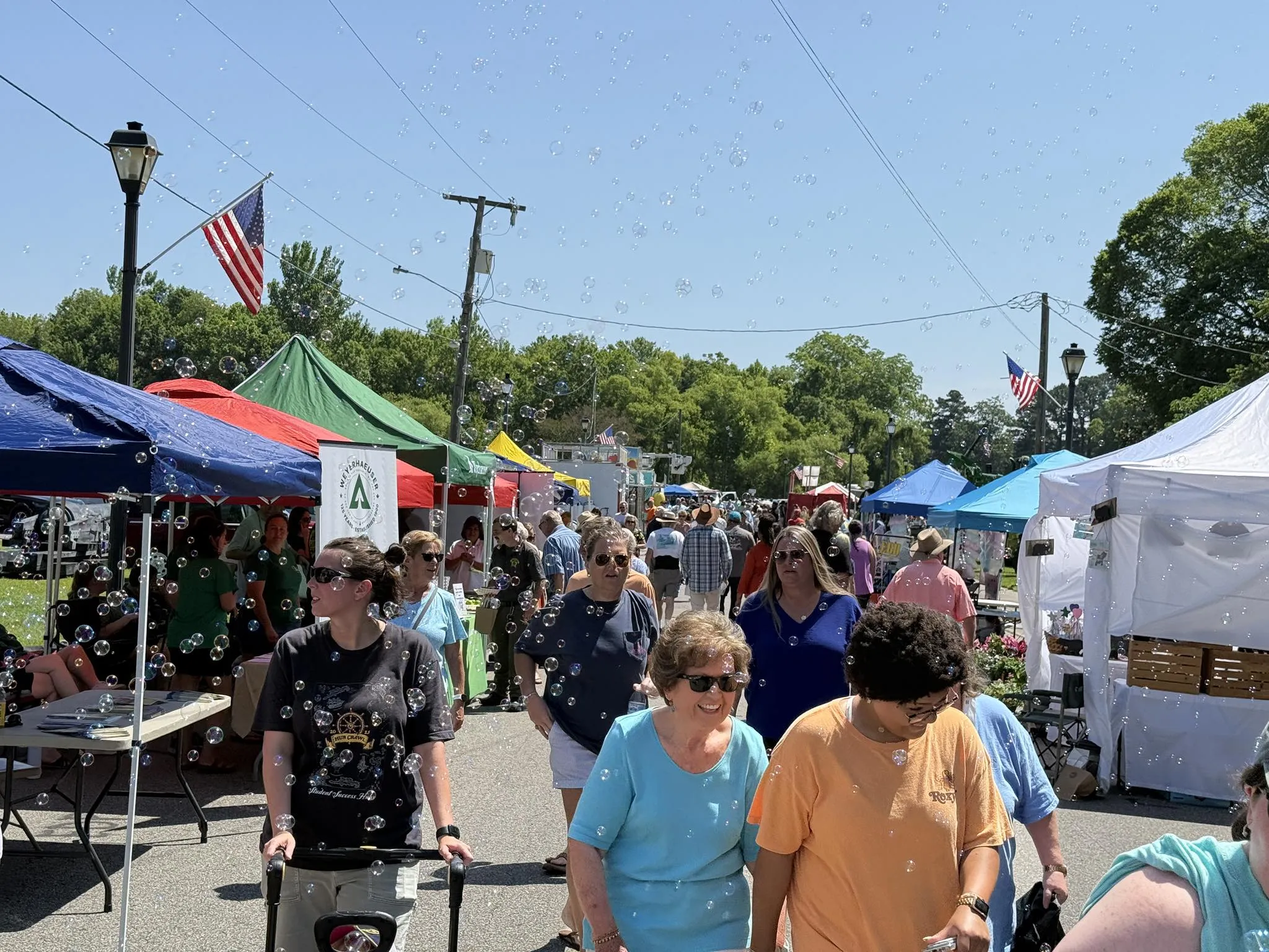 People walk among colorful tents at an outdoor fair under sunny skies, with bubbles floating in the air.