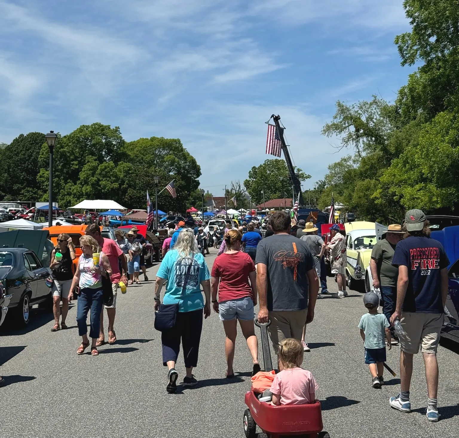 People walk among classic cars at an outdoor car show on a sunny day, with trees and flags in the background.