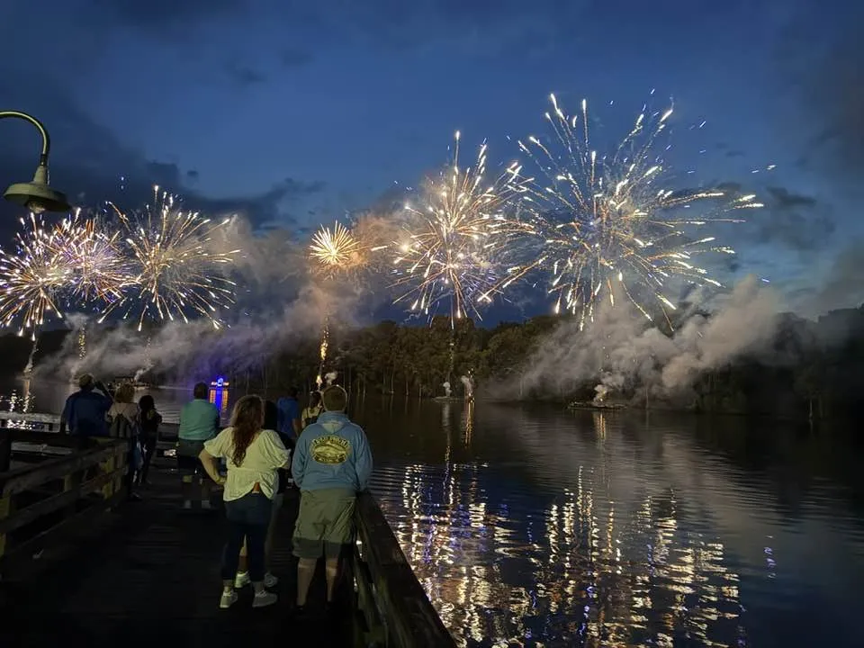 People watch colorful fireworks explode over a lake at night, with reflections shining on the water.