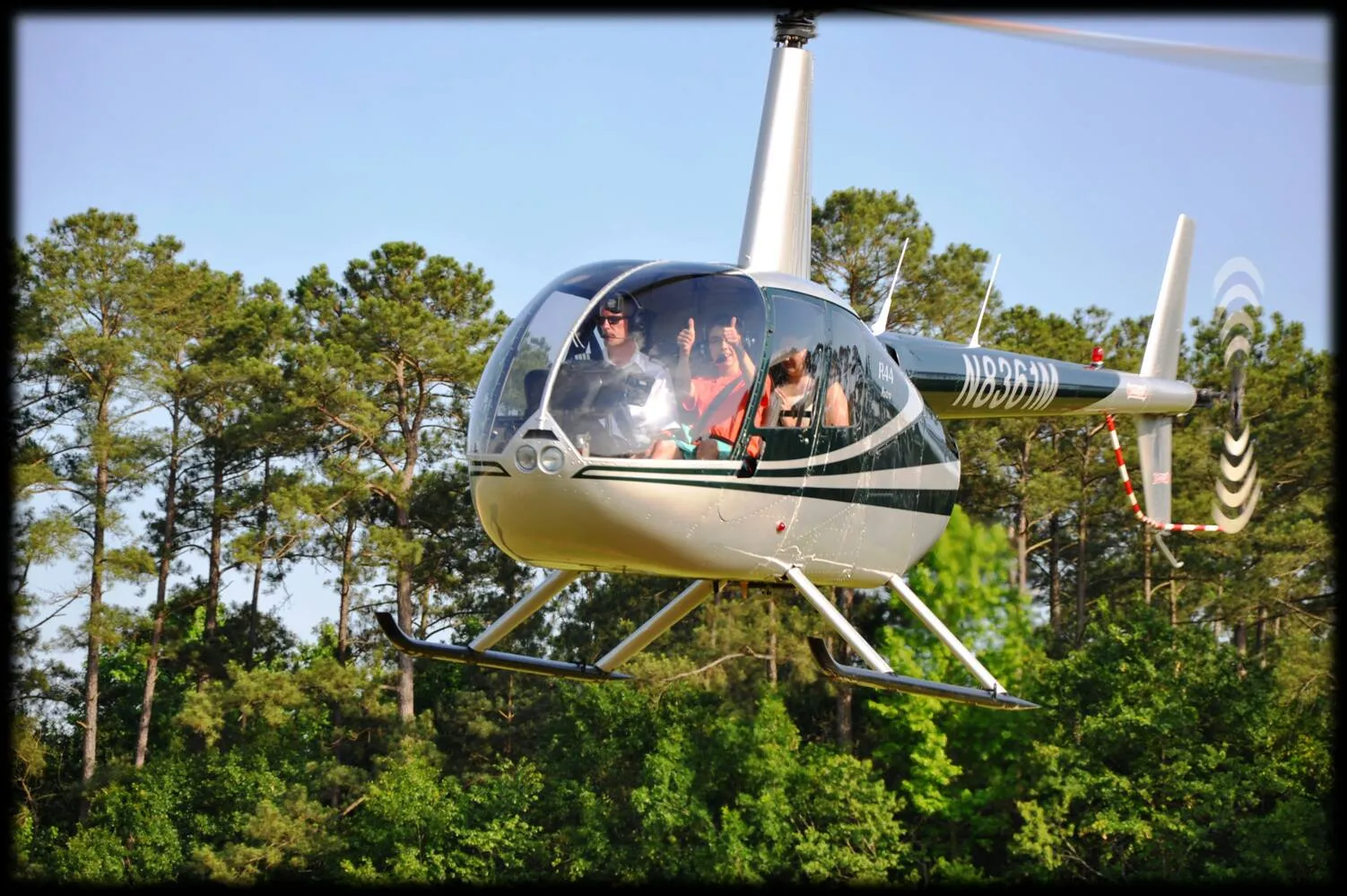 A helicopter with passengers inside flying low above trees on a clear, sunny day.