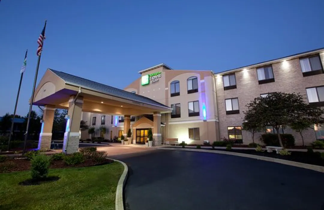 holiday-in-1 Modern hotel building with lit entrance at dusk, American flag, and landscaped garden in front.