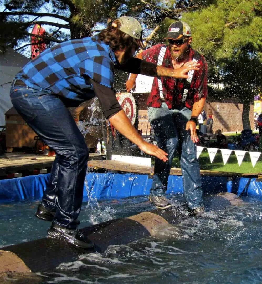 Two men compete in log rolling on water, splashing as they balance on a floating log outdoors.