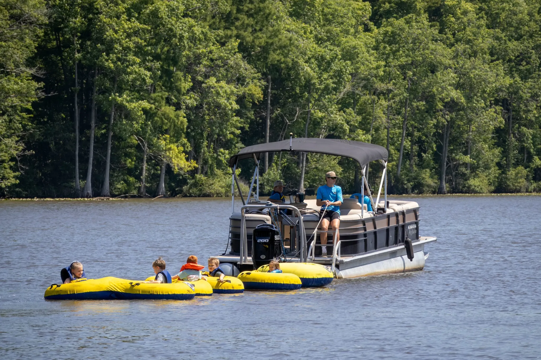 A pontoon boat pulls two yellow inflatable tubes with children on a lake surrounded by trees.