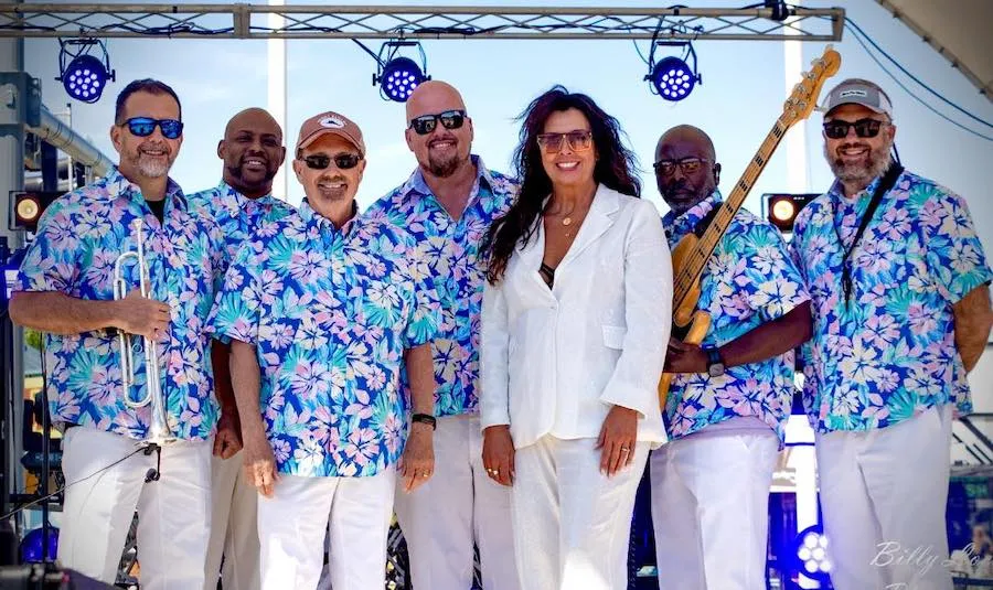 A woman in white stands with six men in blue floral shirts and white pants on an outdoor stage.