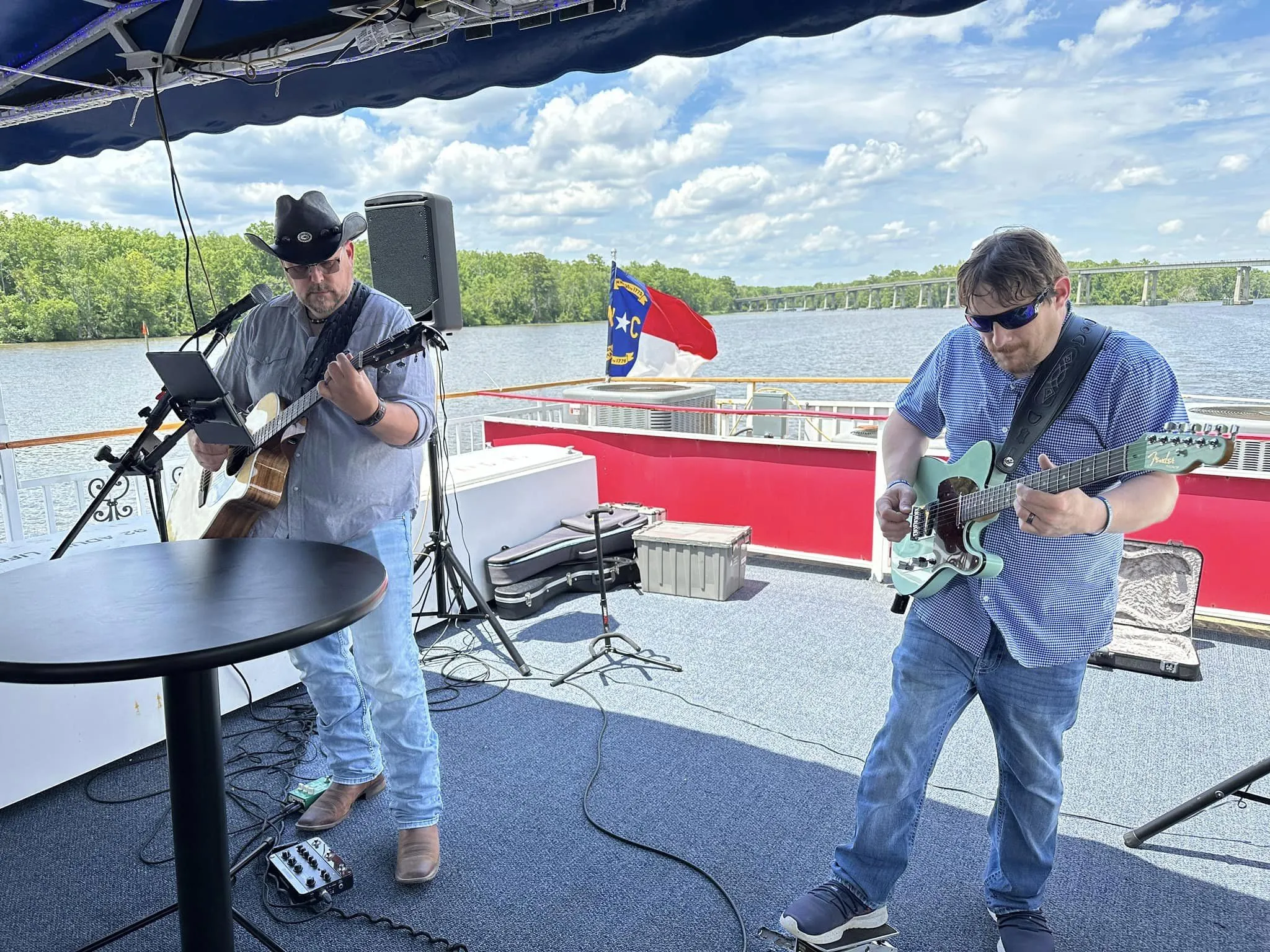 Two men play guitars on a boat deck, with a river, trees, and a flag in the background under a blue sky.