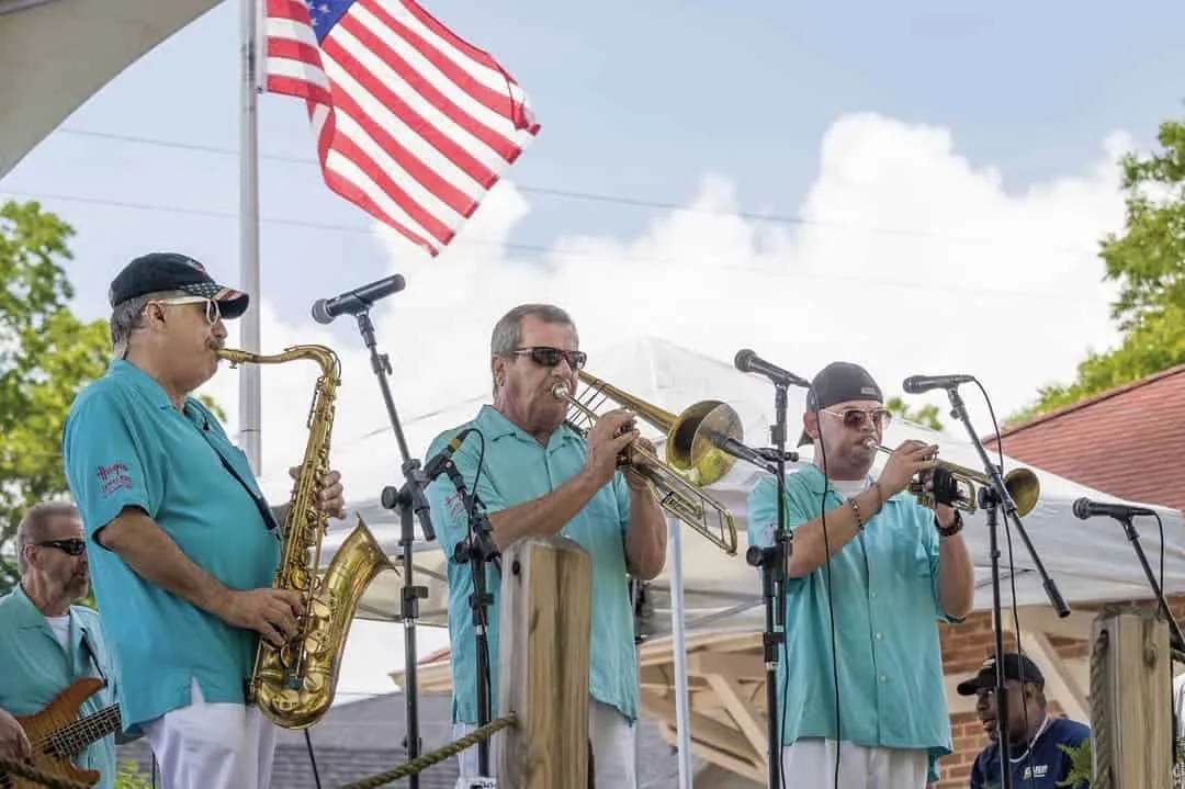 Three musicians in teal shirts play saxophone, trombone, and trumpet on an outdoor stage with an American flag.