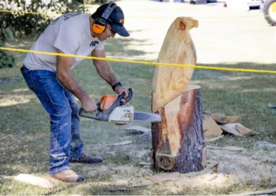 Man wearing ear protection carving a wooden sculpture with a chainsaw outdoors on grass.