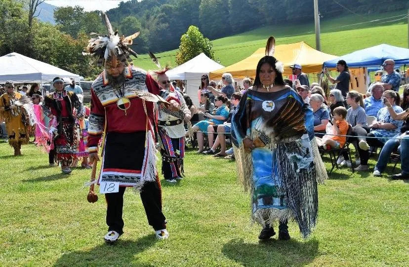 Two people in traditional Native American attire dance outdoors at a cultural event, with a crowd watching.