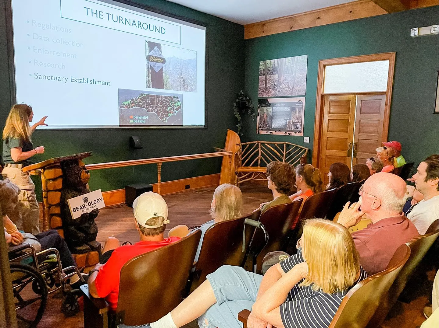 A woman presents a slideshow about bear conservation to an audience seated in a small lecture room.