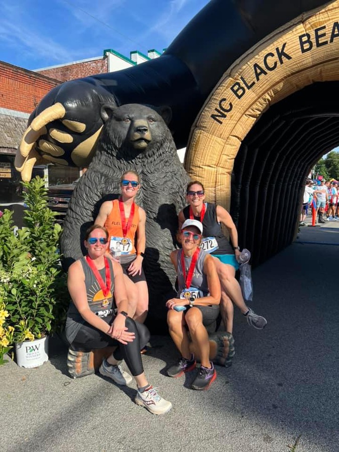 Four runners with medals pose by a bear statue at the NC Black Bear event entrance on a sunny day.