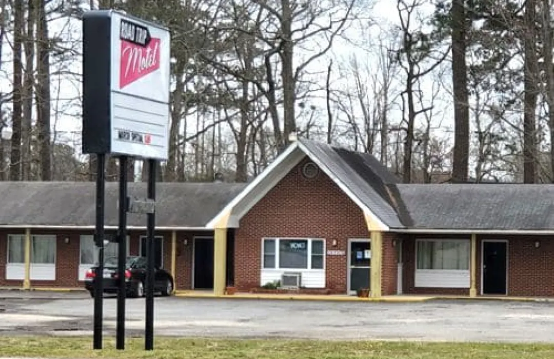roadtrip-1 A small brick roadside motel with a blank sign and several rooms, surrounded by trees and parked cars.