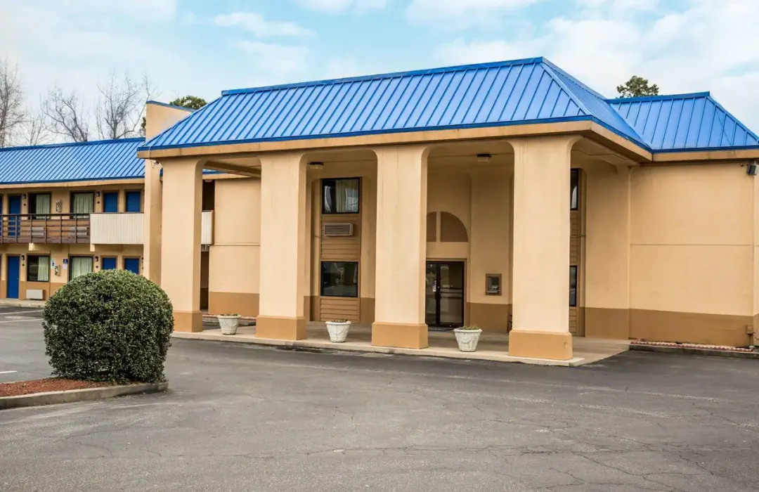 rodeway-inn-1 Beige two-story motel building with blue roof, large columns, and parking lot in front.