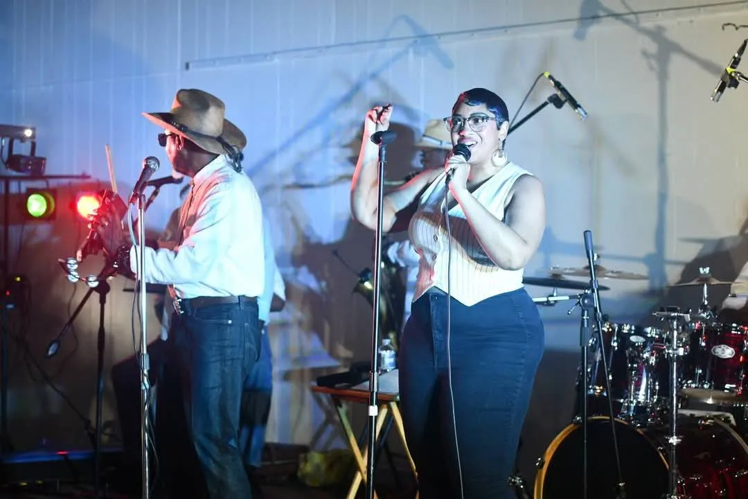 A woman sings into a microphone on stage next to a violinist; drums and instruments are in the background.