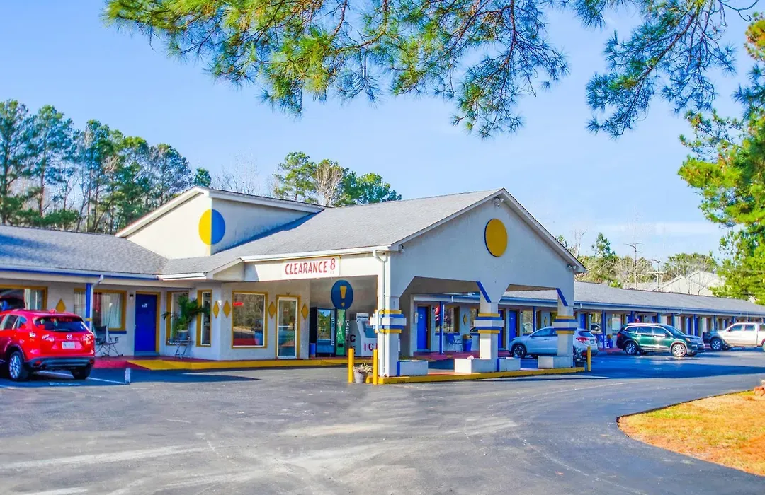 sportsman-inn-1 A roadside motel with parked cars under a clear blue sky, surrounded by trees and pine needles.